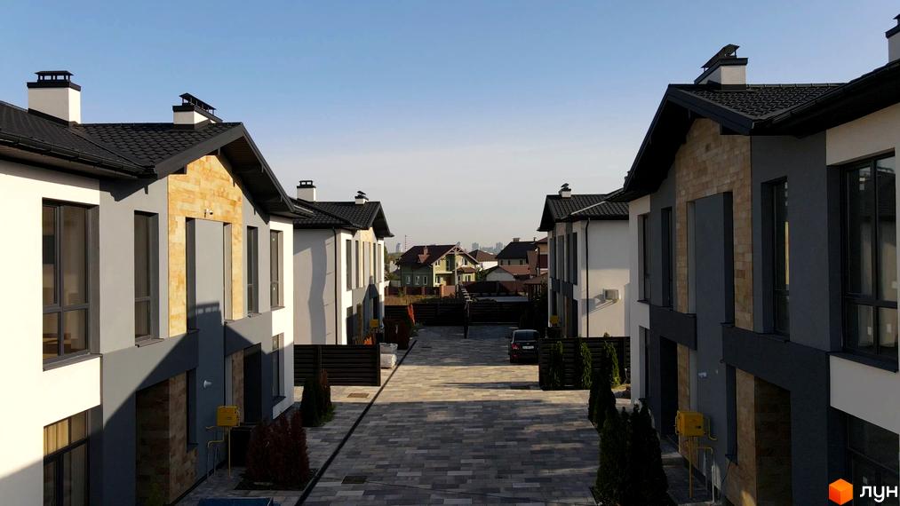 Зображення двоповерхових будинків в ЖК Sandstone Comfort з темною покрівлею та оздобленням фасаду. Бруківка на території та зелені насадження.