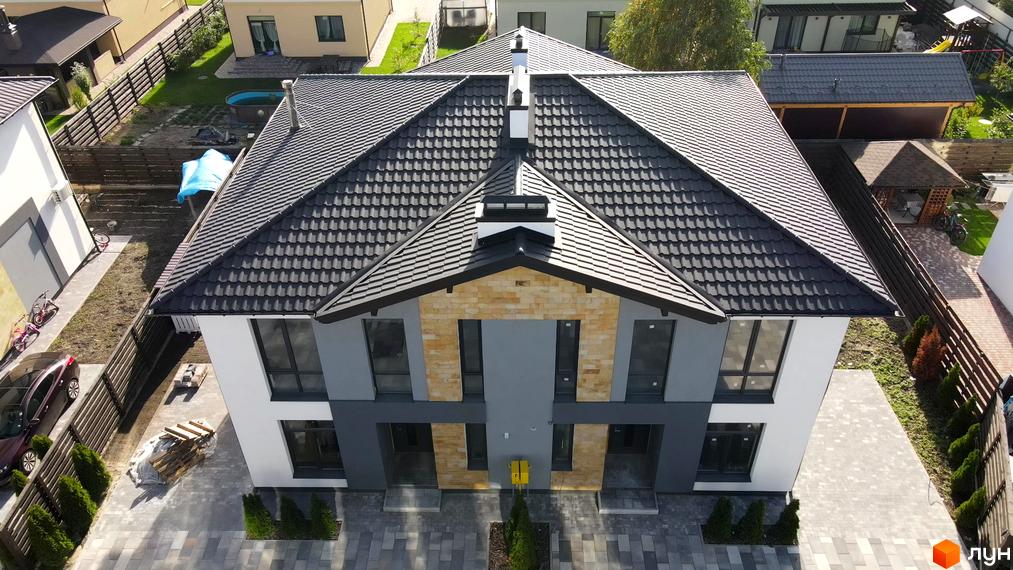 Двоповерховий будинок з темною черепицею на даху. Фасад будинку поєднує сірий, білий та піщаний кольори. Передня територія викладена бруківкою, є зелені насадження.