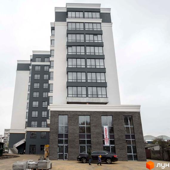 Фасад новобудови ЖК Вільський з великою кількістю вікон та сірими елементами. Поруч припаркований автомобіль та будівельні матеріали.