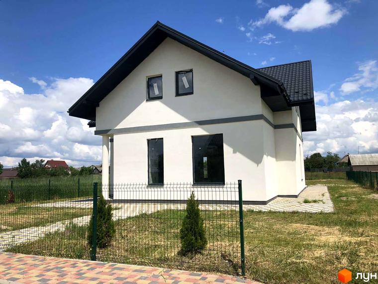 Білий будинок з темним дахом та вікнами. Перед будинком зелений газон, огорожа та бруківка. КМ Лелека - ідеальне місце для життя.
