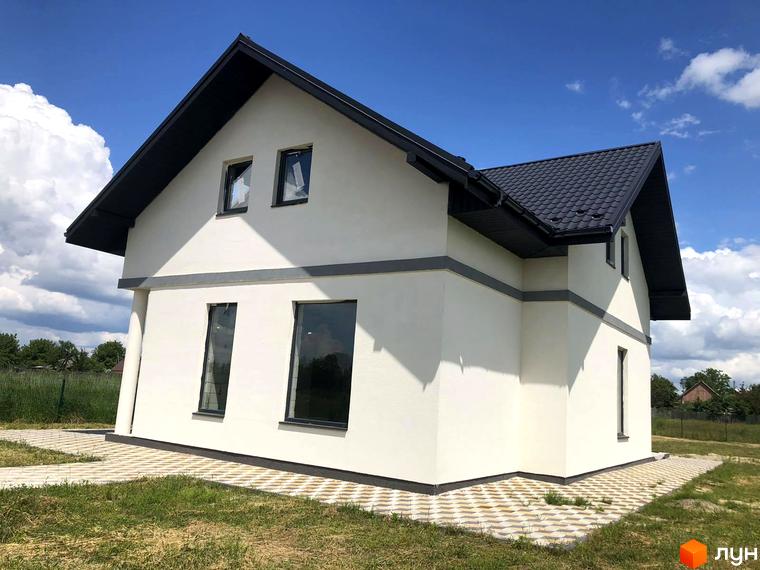 Білий двоповерховий будинок з темним дахом та вікнами. Перед будинком викладена бруківка, навколо газон.