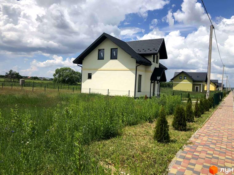 Двоповерховий будинок з темним дахом в Містечку котеджів Лелека. Поруч з будинком розташована доглянута прибудинкова територія з рослинами та тротуаром.