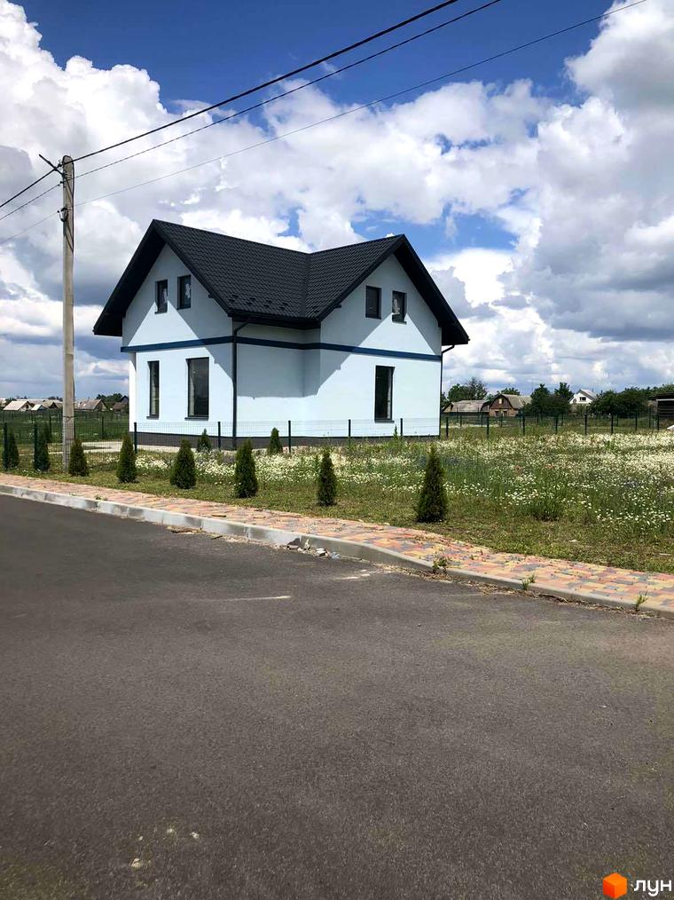 Білий будинок з чорним дахом на тлі блакитного неба з хмарами. Перед будинком розташована зелена трава з квітами, а також дорога.