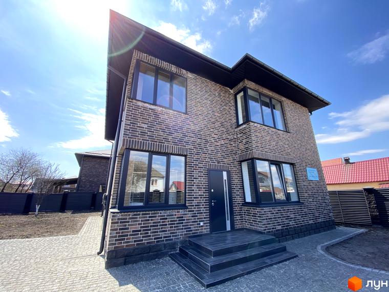Котеджне містечко КМ Весняний: двоповерховий цегляний будинок з темною черепицею та великими вікнами. Поруч вимощена бруківкою доріжка та зелений газон.