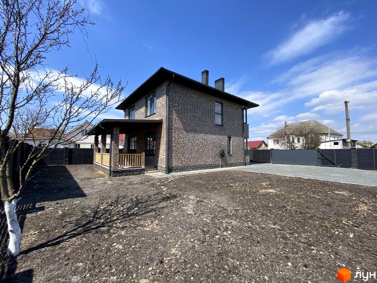 Котеджне містечко КМ Весняний: двоповерховий цегляний будинок з навісом та дерев'яним парканом. Прибудинкова територія з деревами та парканом.