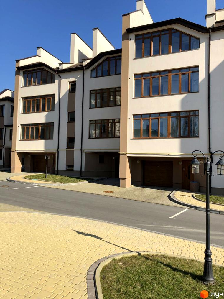 Фасад житлового комплексу світлого кольору з коричневими вікнами та гаражами. Прибудинкова територія має жовту бруківку та зелені газони.