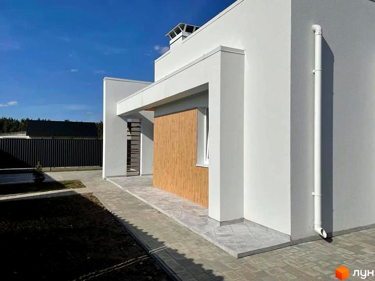 Зовнішній вигляд будинку котеджного містечка КМ Лісне з білою штукатуркою та дерев'яними елементами. Поруч вимощені доріжки та зелена ділянка.