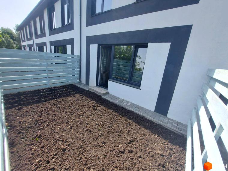 Таунхаус Sanny House з біло-сірим фасадом та великим вікном, перед яким розташована ділянка з чорноземом та дерев'яною огорожею.