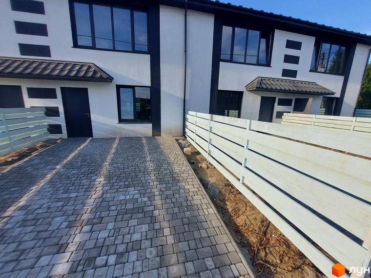 Зовнішній вигляд таунхауса Sanny House з сірим брукованим подвір'ям та білим парканом. Новобудова з чорними вікнами та дверима.