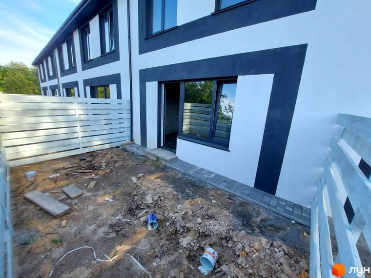 Таунхаус Sanny House з біло-темно-сірим фасадом та вікнами. На прибудинковій території видно паркан, будівельні матеріали та облаштування території.