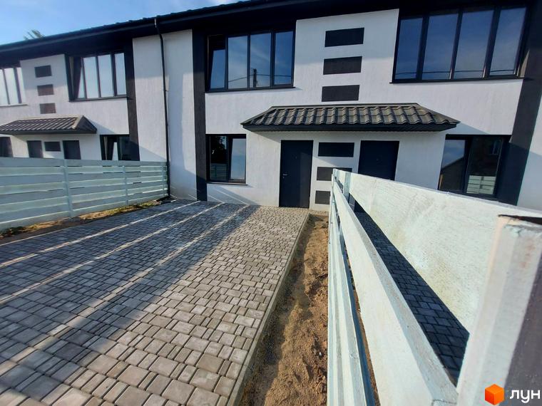 Таунхаус Sanny House з біло-чорним фасадом та великими вікнами. Повністю вимощена бруківкою територія перед будинком, огороджена дерев'яним парканом.