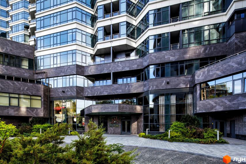 Фасад Житлового комплексу PecherSKY з темно-сірим облицюванням та великими скляними вікнами. Перед входом розташовані доріжки з бруківки та доглянуті зелені насадження.