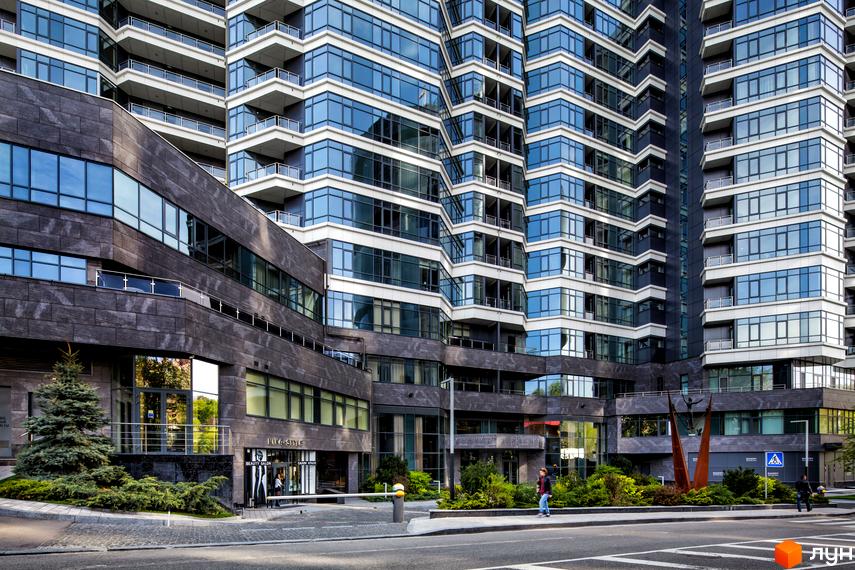 Багатоповерховий житловий комплекс PecherSKY зі скляними фасадами та темною кам'яною обробкою. Навколо будівлі розташовані зелені насадження та пішохідна зона.