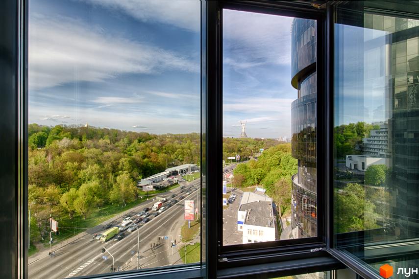 Житловий комплекс PecherSKY: висотний будинок з вигнутим скляним фасадом, що виходить на зелений парк, жваву дорогу та міський пейзаж з пам'ятником.