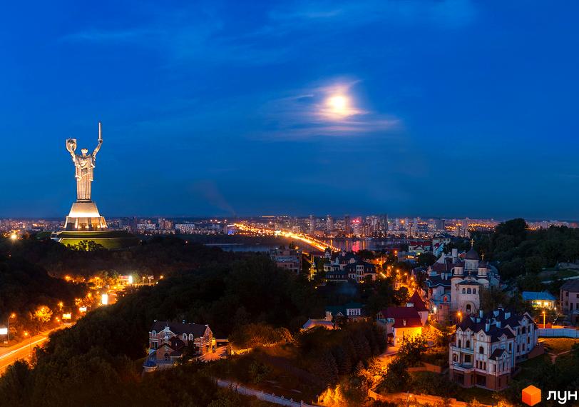 Вид на житловий комплекс PecherSKY з вечірнім освітленням, поруч зелені насадження та забудова. На задньому плані місто з вогнями.