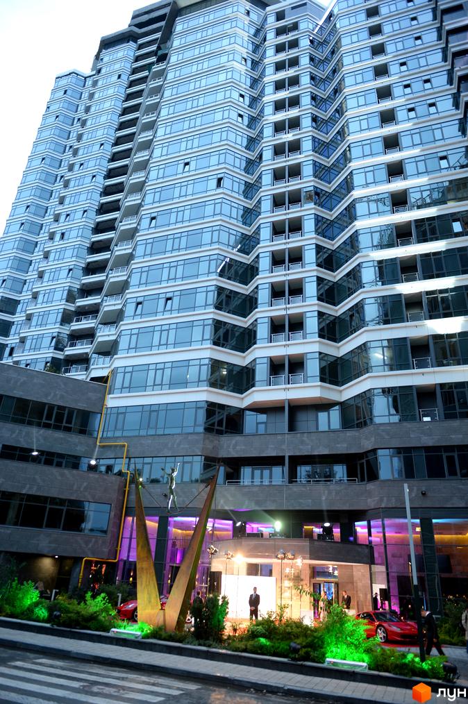 Високий будинок Житлового комплексу PecherSKY зі ступінчастим скляним фасадом. Перед будівлею зелені насадження, червоний спортивний автомобіль та скульптура.