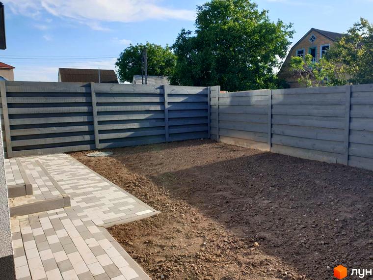 Сірий дерев'яний паркан навколо прибудинкової території житлового комплексу на вулиці Шкільна, 2. На території видно вимощену плиткою доріжку та ділянку з ґрунтом.