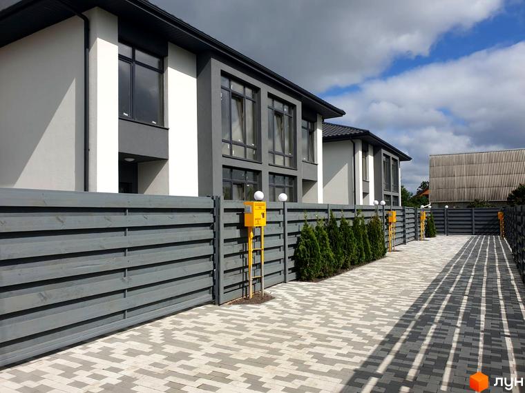 Зовнішній вигляд будинків житлового комплексу на вул. Шкільна, 2 з сірим дерев'яним парканом та вимощеною територією. Сучасна архітектура та доглянута прибудинкова територія.