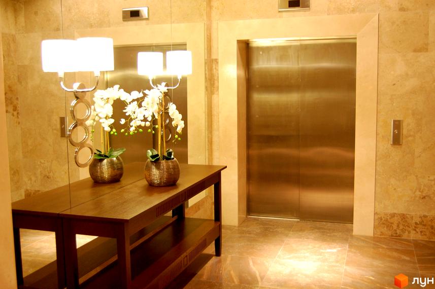 Вестибюль Житлового комплексу PecherSKY з двома ліфтами та оздобленням стін світлою мармуровою плиткою. У холі розташований дерев'яний стіл з декоративними орхідеями.