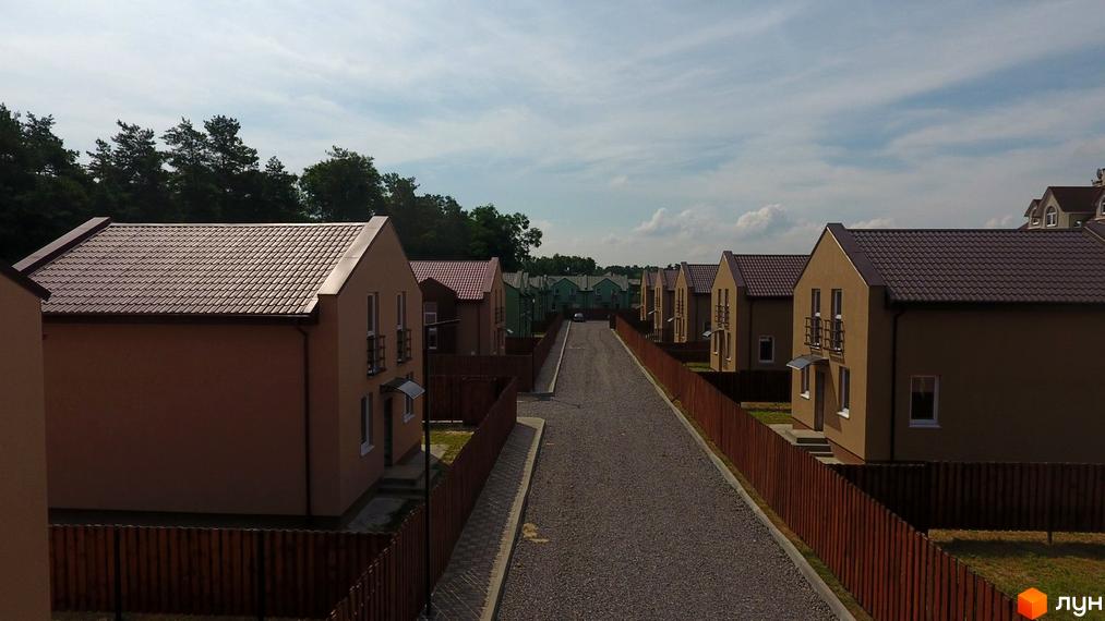 Котеджне містечко Sunny Village: двоповерхові будинки зі світлими фасадами та коричневими дахами. Вулиця з гравійним покриттям та зелена прибудинкова територія.