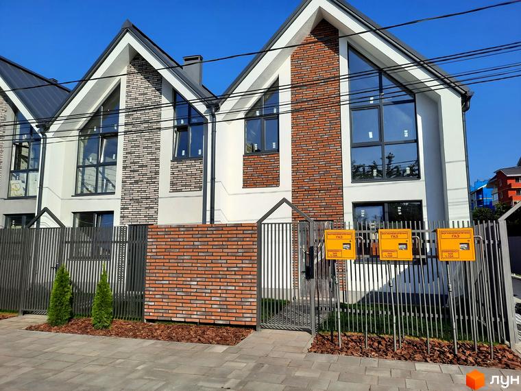 Зовнішній вигляд будинків у Котеджному містечку Villa Gatne з цегляними вставками та великими вікнами. Перед будинками металева огорожа та зелені насадження.