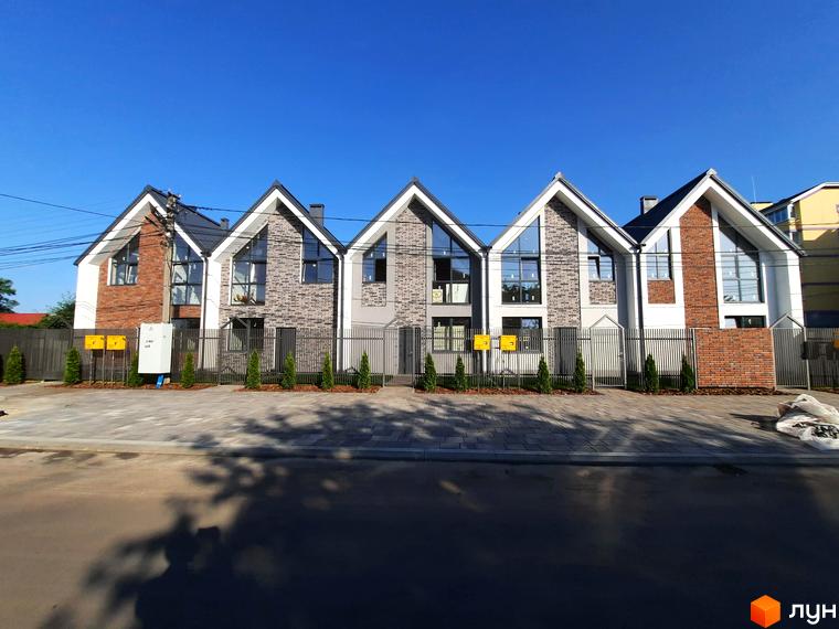 Зовнішній вигляд будинків у Котеджному містечку Villa Gatne з сірого та червоного цегли, перед будинками висаджені туї та встановлена огорожа.