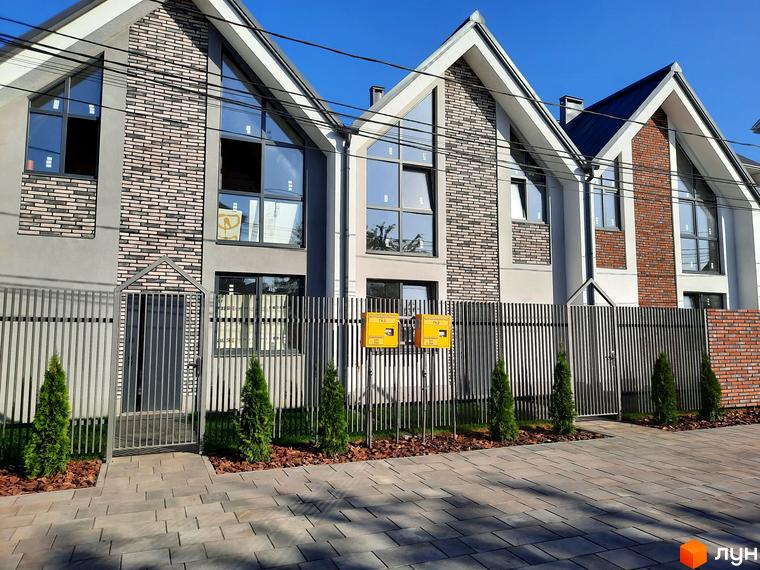 Зображення фасаду та прибудинкової території Котеджного містечка Villa Gatne. Вигляд новобудови з сірим парканом та зеленими насадженнями.