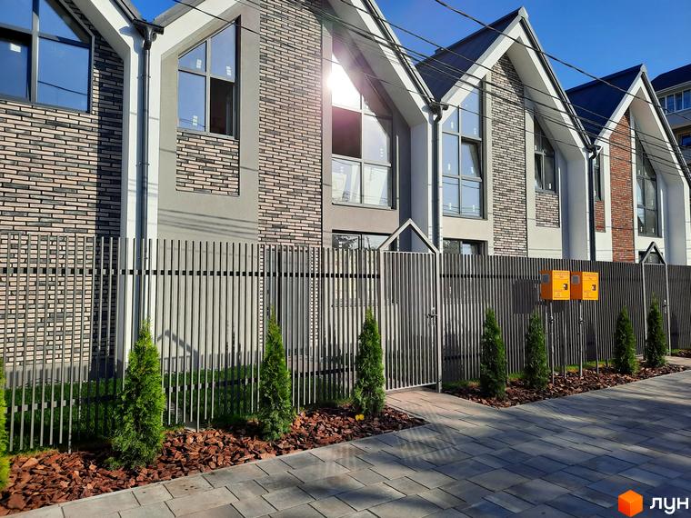 Зовнішній вигляд Котеджного містечка Villa Gatne з сірою огорожею та невеликими туями вздовж тротуару.