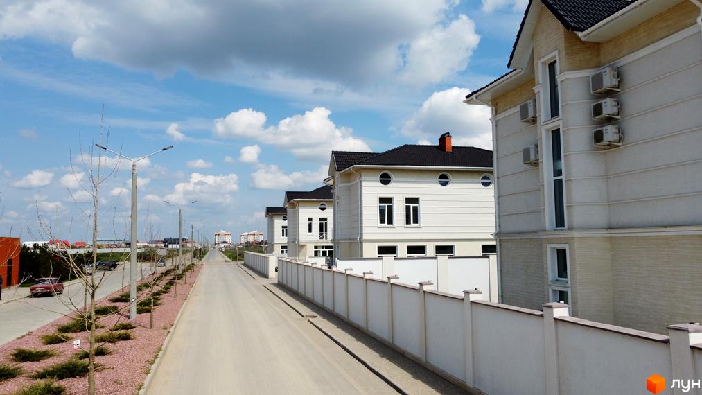 Білі двоповерхові будинки Котеджного містечка Дайберг з темними дахами та кондиціонерами на фасаді. Вздовж дороги розташовані ліхтарі та молоді дерева.