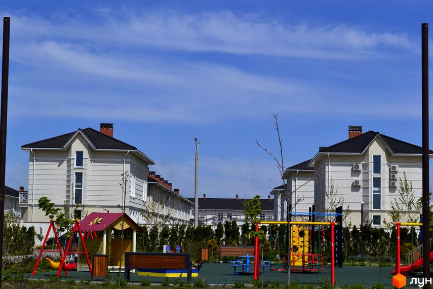 Котеджне містечко Дайберг: світлі будинки з темними дахами та дитячий майданчик. На передньому плані розташований ігровий майданчик з гойдалками та лавками.