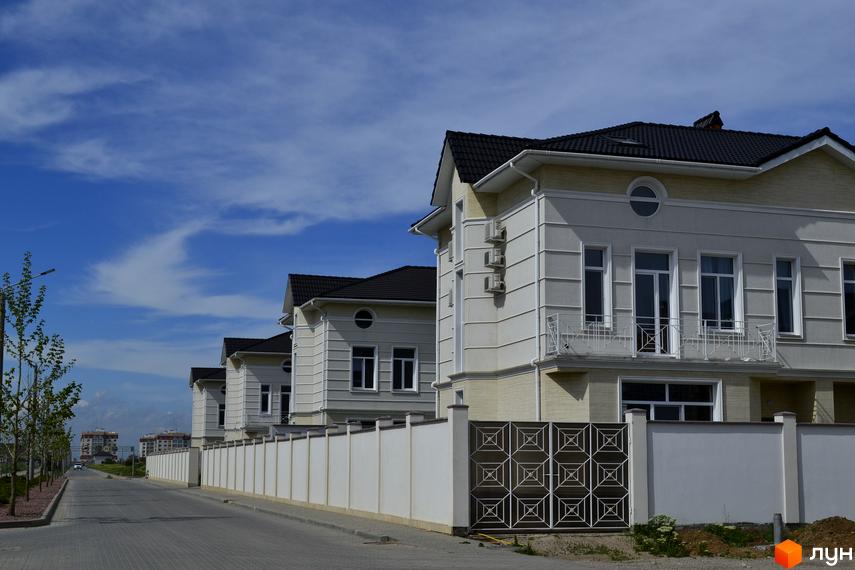 Котеджне містечко Дайберг: світлі двоповерхові котеджі з темними дахами та балконами. Дорога, білий паркан та молоді дерева біля будинків.