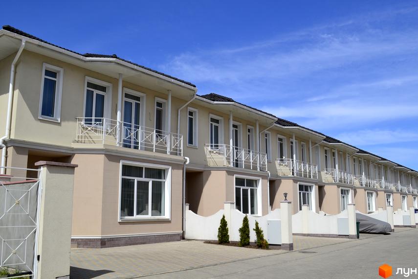 Ряд бежевих котеджів Котеджного містечка Дайберг з білими балконами та вікнами. Перед будинками бруківка та зелені насадження.