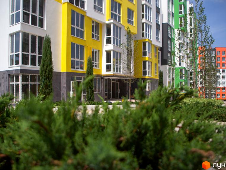 Житловий комплекс Синергія-3+ з яскравими фасадами жовтого, зеленого та помаранчевого кольорів. На передньому плані зелені кущі та дерева.
