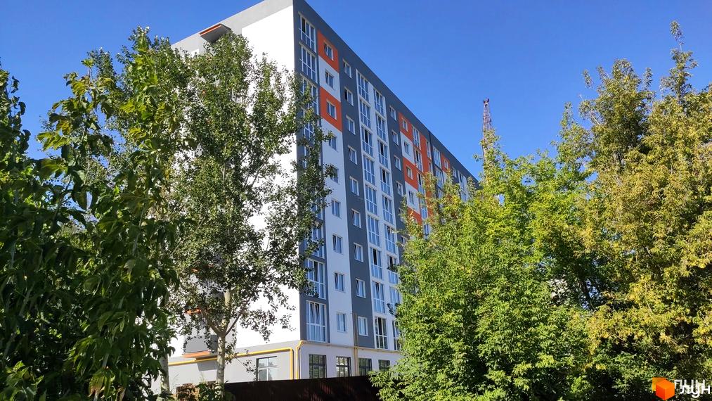 Зовнішній вигляд житлового комплексу на вул. Піщана, 1б, з фасадом сірого, білого та помаранчевого кольорів. Навколо будівлі розташовані зелені дерева.