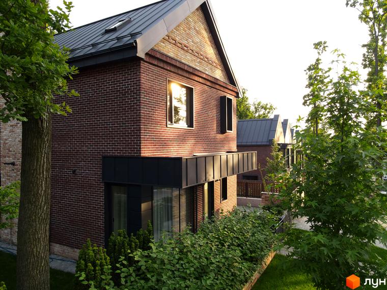 Фасад цегляного будинку з темним металевим дахом та вікнами в Котеджному містечку Oak Grove Town. Прибудинкова територія озеленена деревами та кущами.