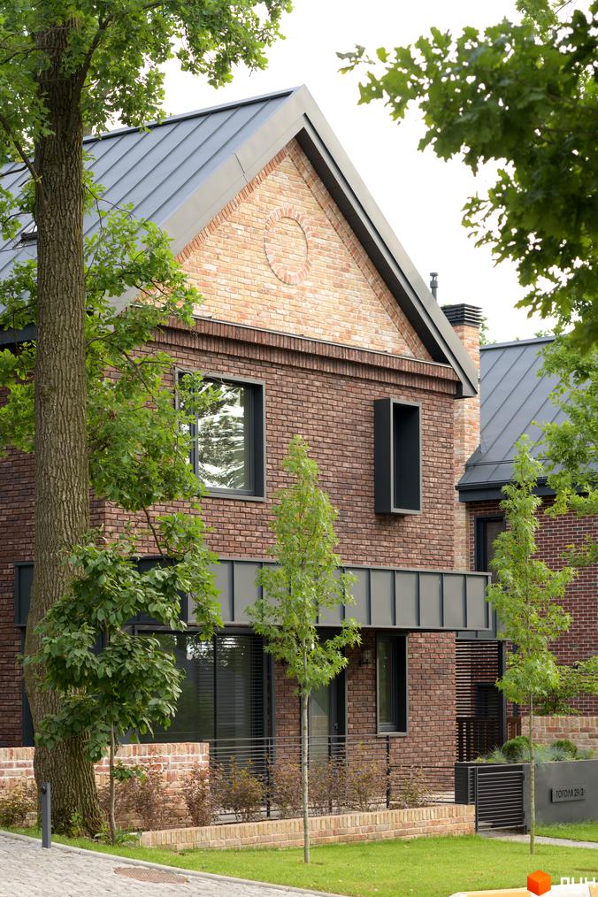 Цегляний будинок в КМ Oak Grove Town з темною покрівлею та великими вікнами. На прибудинковій території газон, дерева та низький паркан.