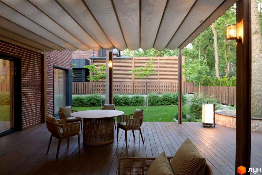 Тераса котеджу Котеджне містечко Oak Grove Town з червоної цегли, дерев'яним настилом та обідньою зоною. Зелений газон та дерев'яний паркан оточують будинок.