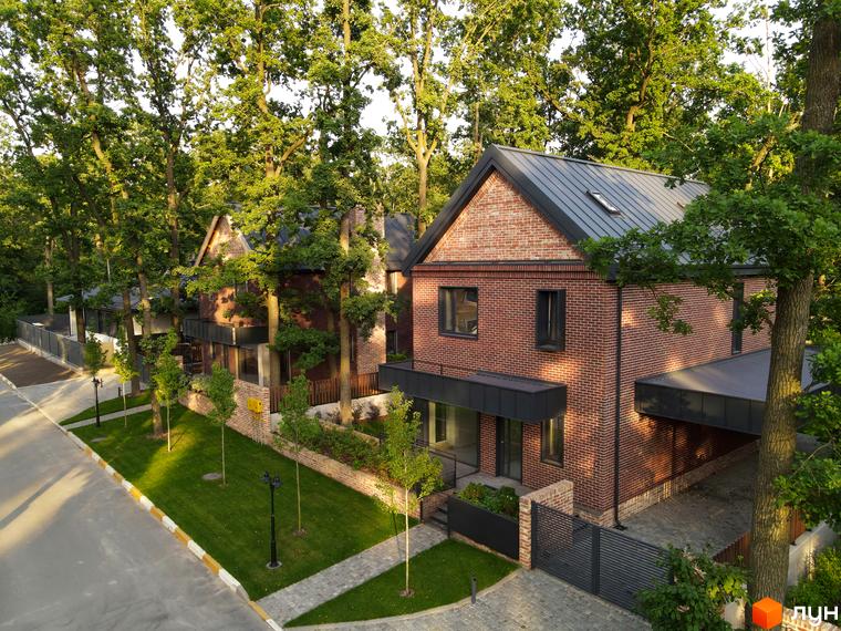 Будинки Котеджного містечка Oak Grove Town з червоної цегли та темними дахами, розташовані серед густих дерев. Прибудинкова територія має доріжки та доглянуті газони.