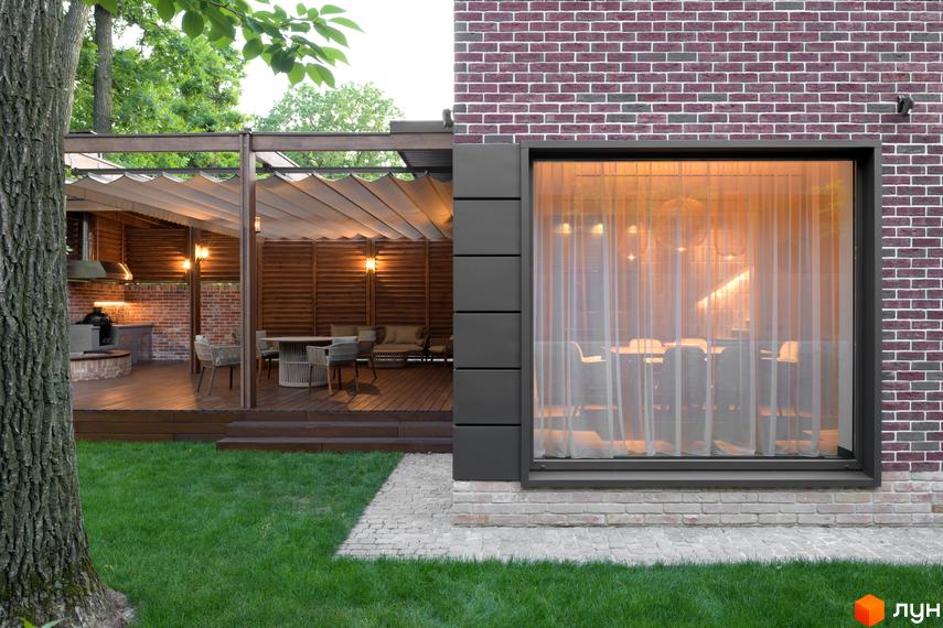Зовнішній вигляд будинку з цегляною кладкою та великим вікном. Поруч розташована тераса з дерев'яними елементами та зелена галявина з деревом.