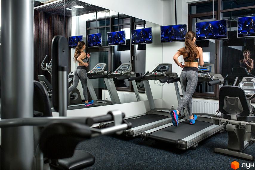 Житловий комплекс Skyline: жінка тренується на біговій доріжці у сучасному спортзалі з великим дзеркалом та телевізорами.
