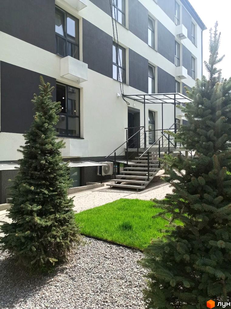 Фасад будинку з чорно-білим оздобленням та сходами до входу. Навколо будинку зелений газон та хвойні дерева.