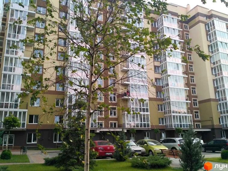 Багатоповерховий будинок Житлового комплексу Oliva з балконами та великими вікнами. На передньому плані видно зелені насадження та припарковані автомобілі.