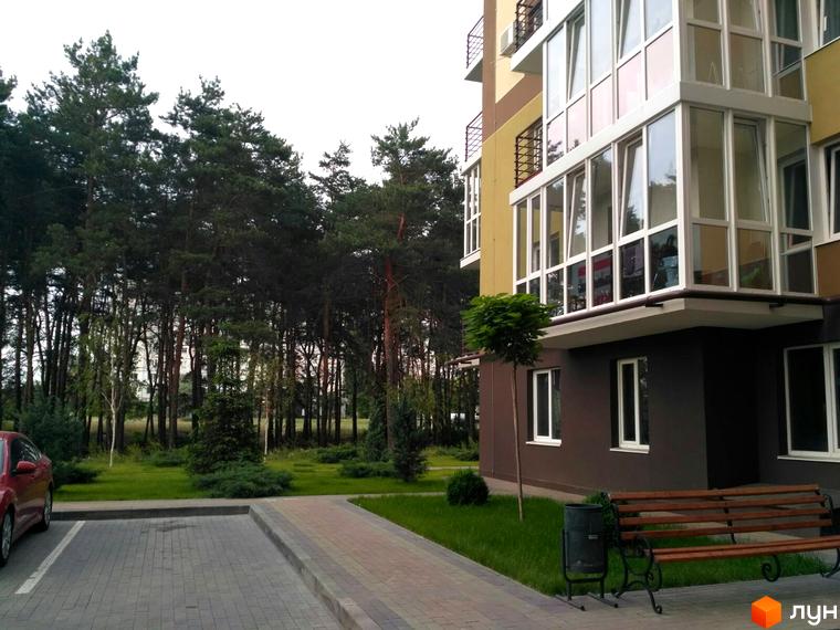 Фасад будинку Житлового комплексу Oliva жовто-коричневий, з великими вікнами. Прибудинкова територія включає зелені газони, лавки та парковку біля лісу.