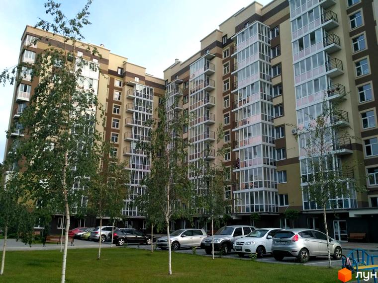 Житловий комплекс Oliva – це багатоповерхові будинки з панорамним склінням та балконами, розташовані біля зеленої зони з деревами та місцями для паркування.