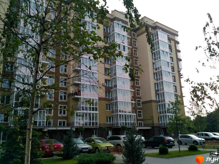 Новобудова Oliva: багатоповерховий будинок світлих тонів із заскленими балконами. Прибудинкова територія має зелені насадження та місця для паркування.