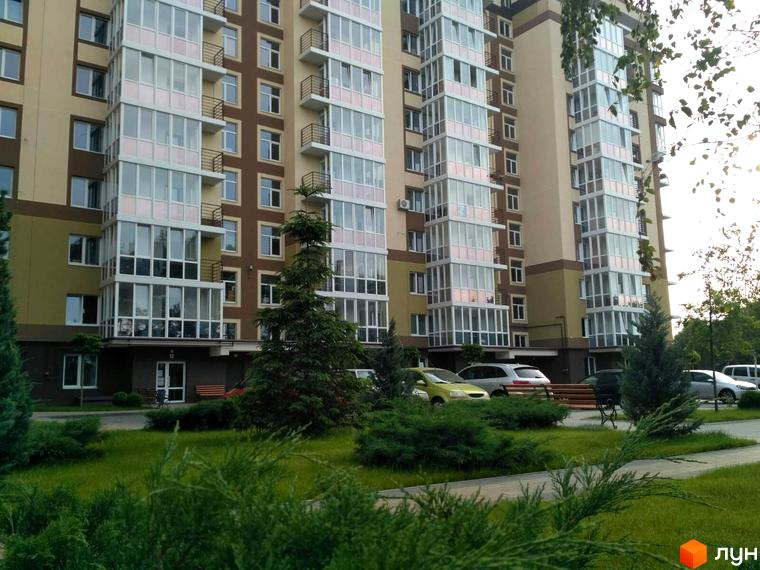 Житловий комплекс Oliva: багатоповерховий будинок бежевого та коричневого кольорів зі скляними балконами. На території зелений газон, кущі, дерева та припарковані авто.