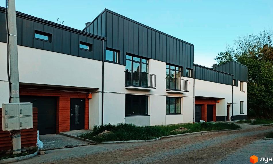 Зовнішній вигляд котеджного містечка КМ Медовий: фасади будинків з темним дахом та білими стінами, балкони, гаражні ворота. Прибудинкова територія з бруківкою та зеленню.