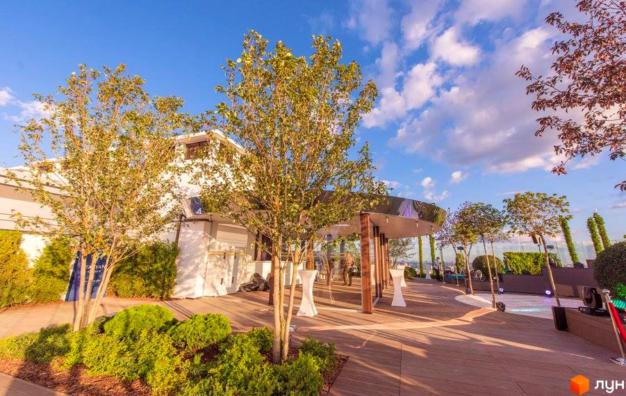 Royal Tower - Будівельний проєкт з білими фасадами та терасою з дерев'яним покриттям. На території багато зелені, дерева та кущі.