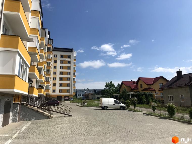 Зображення фасаду та прибудинкової території Житлового комплексу Світанковий. Багатоповерховий будинок з жовтими балконами, подвір'я з тротуарною плиткою.