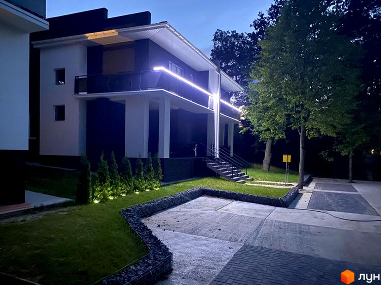 Котеджне містечко КМ Терещенські Озера: двоповерховий будинок з підсвіченим балконом та акуратним ландшафтом з ялинок та газону.
